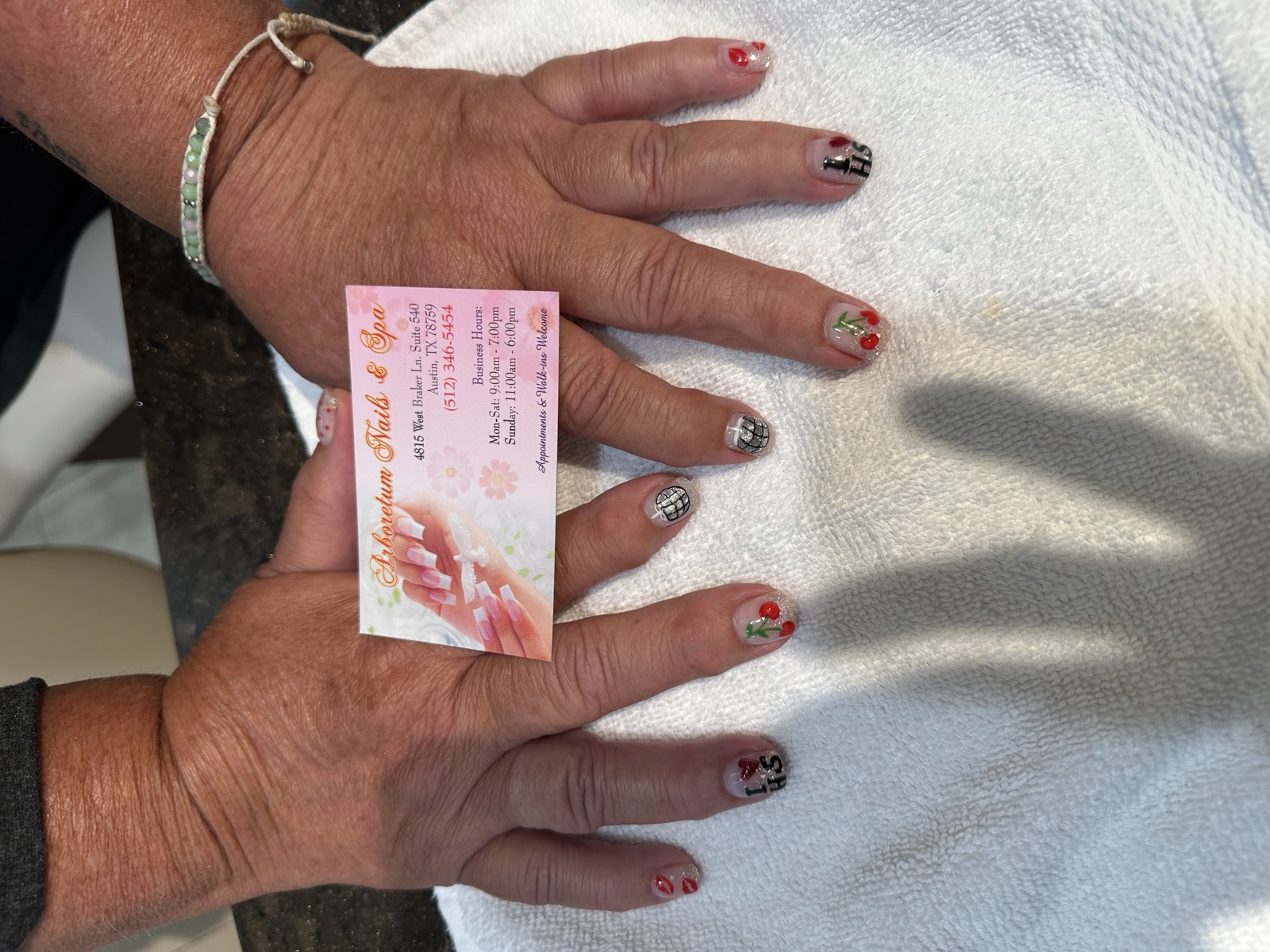 Clear nails with cherry, disco-ball, and letter nail art holding an Arboretum Nails business card